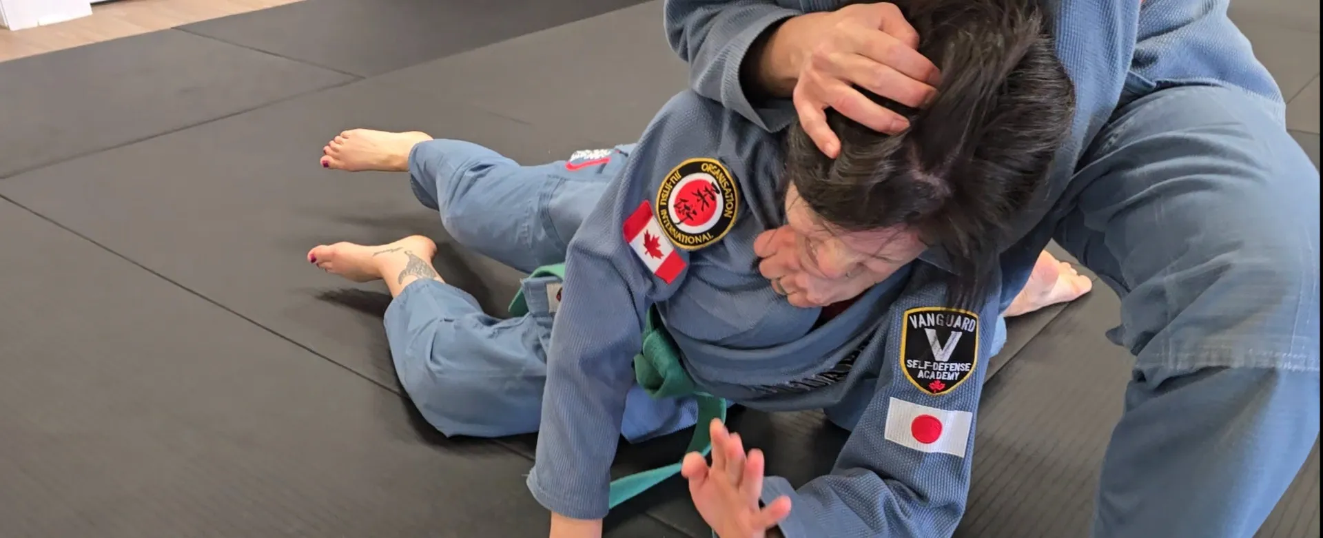 Two adult women in blue gis practicing a controlled ground-defense technique on the Vanguard mats
