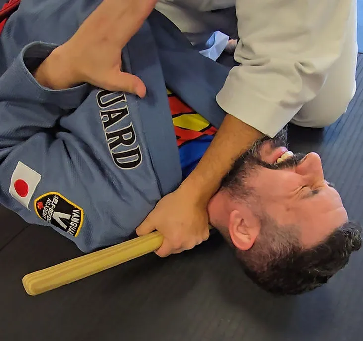 Adult Vanguard student in a blue gi practicing a controlled ground-defense drill with a training stick visible